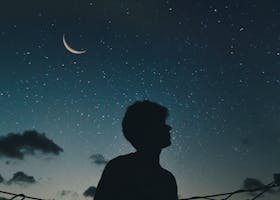 A lone silhouette stands beneath a crescent moon and starry sky, evoking a sense of solitude and wonder.