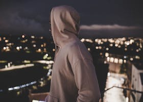 A person in a hoodie overlooks a blurry cityscape at night, evoking mystery and solitude.